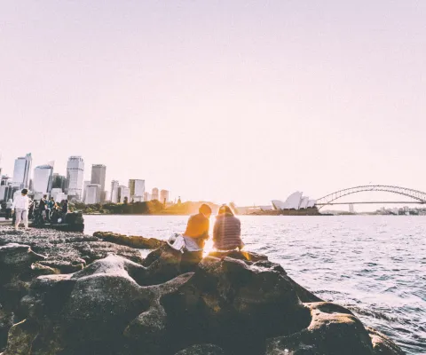 Lidé odpočívají na skalách u sydneyského přístavu, v dálce panorama města